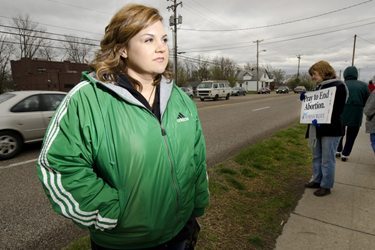 [Photo: Abby Johnson stands on a sidewalk in a green jacket. In the background, a woman holds a sign saying: Pray to End Abortion.]