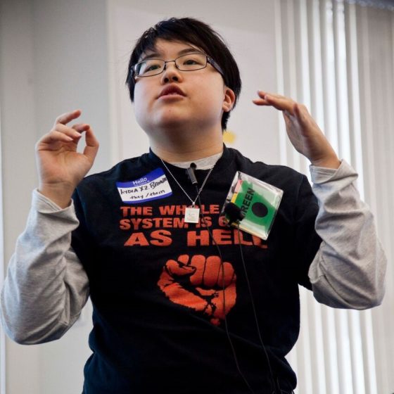 [Photo: Lydia Brown, a young East Asian person, giving a short talk at the Disability Intersectionality Summit in Boston, Massachusetts, November 2016. They are gesturing with both hands. Their t-shirt says 