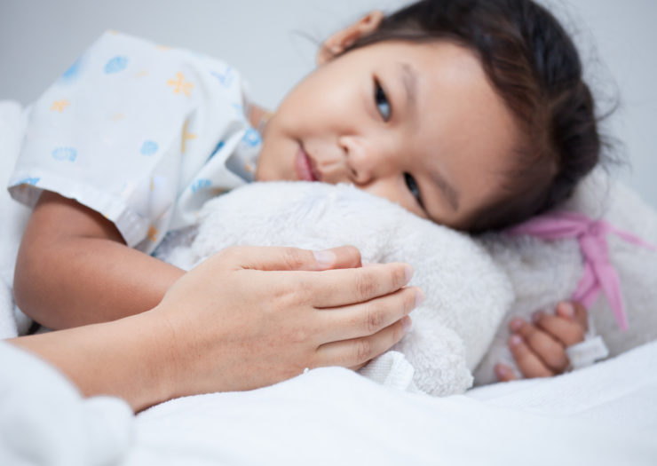 [Photo: A young Asian girl is lying in a bed. She is resting her head on a white stuffed animal with a pink ribbon. A person, who is not seen in the picture, has their hand touching one of the girl's hands.]