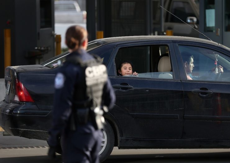 [Photo: A Customs and Border Patrol Officer looks on as a car drives through the San Ysidro Port of Entry. A child in the car looks out the window.]