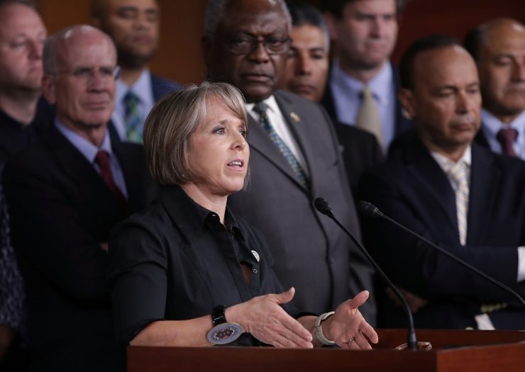 [Photo: Michelle Lujan Grisham speaks at a podium with dozens of of Democratic members of the House of Representatives standing behind her.]