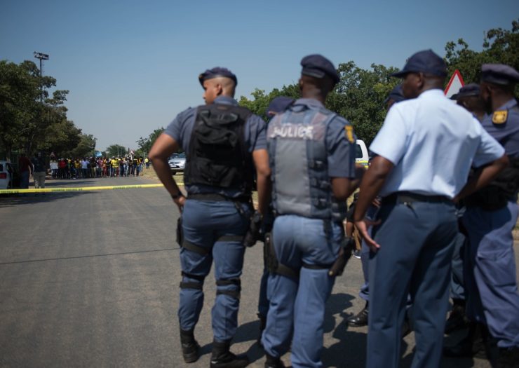 [Photo: South African police officers stand guard in front of a crowd of people.]