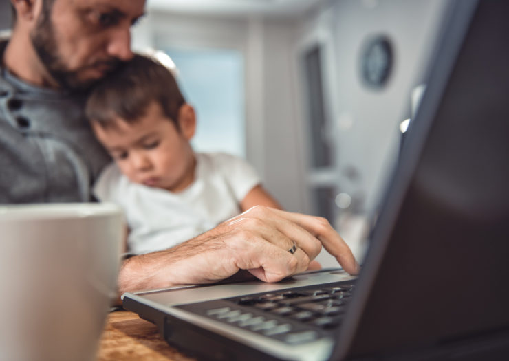 [A man holds his child on his lap while he works on his laptop.]