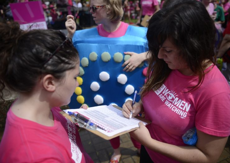 [Photo: A Planned Parenthood activist writes down information on a clipboard while speaking to people in a rally.]