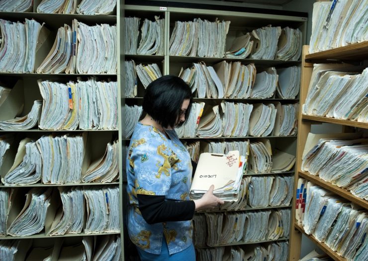 [Photo: A staff member holds files in a medical records room of a hospital.]