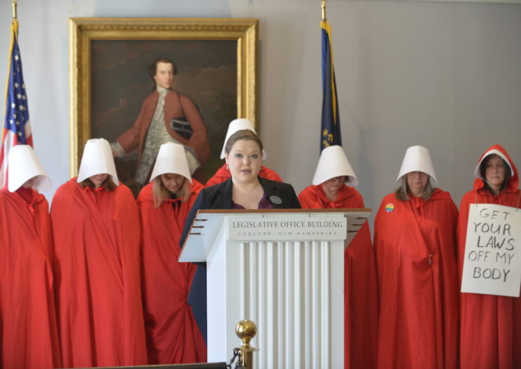 [Photo: Group of women in red 'Handmaid's Tale' costumes stand at press conference behind woman speaking at podium]