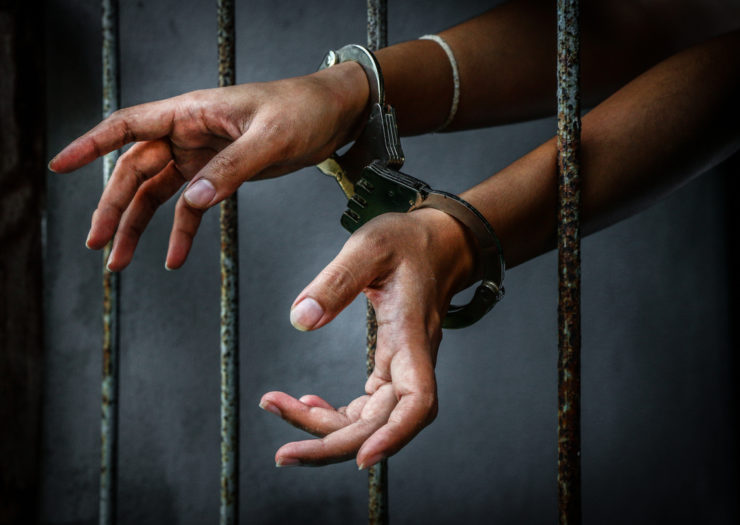 [Photo: A prisoner's hands reach around the bars of their cell, connected by handcuffs.]
