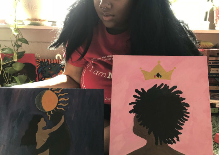 [Photo: A Black woman sits holding two vibrant paintings, one dark blue, one pink. Only the bottom half of her face is visible.]