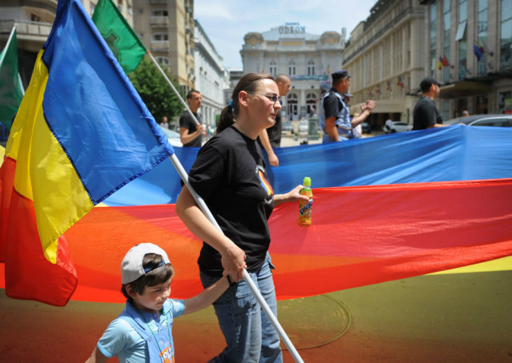 [Photo: A mother holds her child's hand in front of a rainbow flag.]