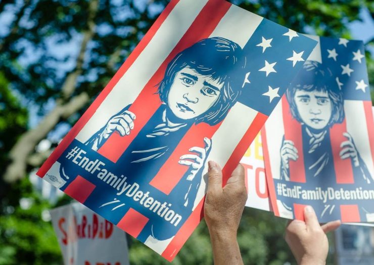 [Photo: People hold illustrations of a child behind the United States flag, the red stripes are like jail bars.]