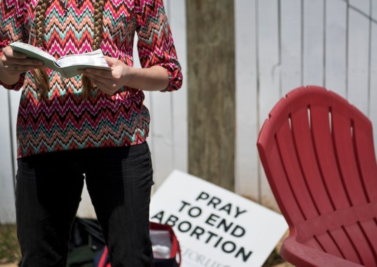 [Photo: An anti-choice activist waits outside of a clinic while reading a bible. The sign behind the activist is 