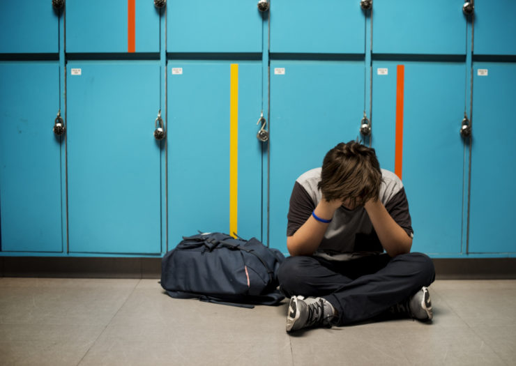 [Photo: A sad high school student sits near lockers with their head in their hands]