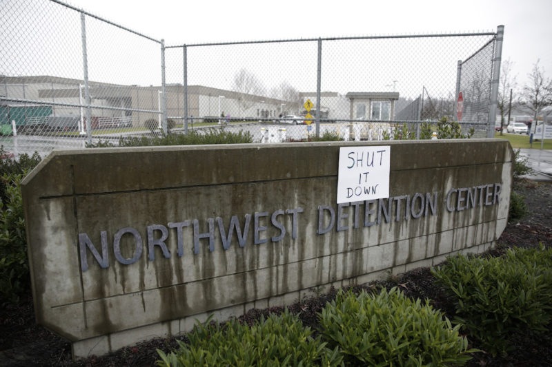 [Photo: Exterior shot of the Northwest Detention Center sign, on which someone posted a paper that reads,