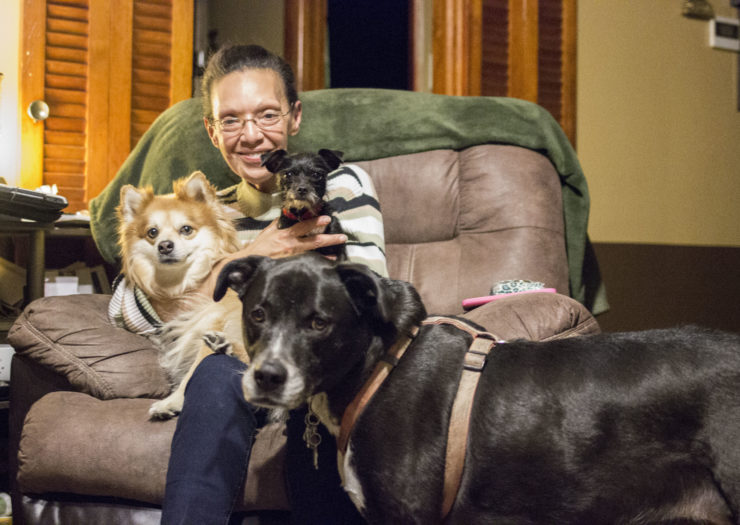 [Photo: Maritza Lopez sits in a recliner, smiling and holding her three dogs.]