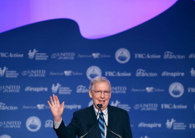 [Photo: Mitch MCConnell waving in front of blue background.]