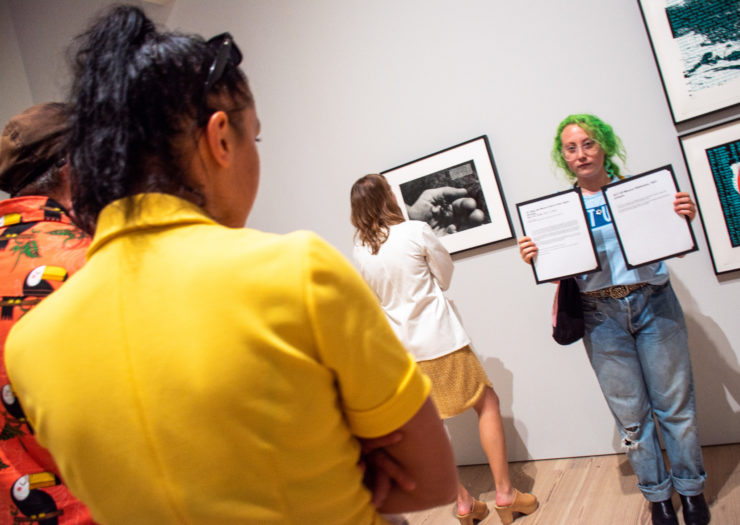 [PHOTO: A young person with neon green hair holds framed papers in an art gallery.]