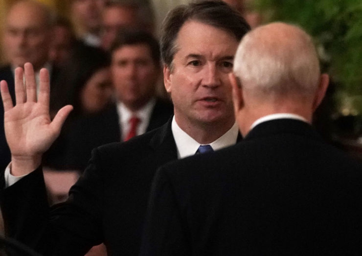 [Photo: Nondescript white man in a suit holds up his hand and stands in front of balding white man, also in a dark suit.]