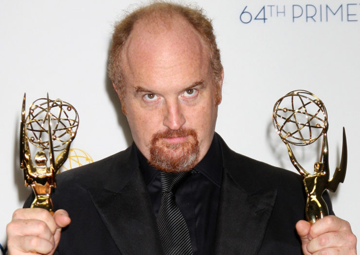 [Photo: Balding white man in a black suit holds a gold award statues in each hand.]
