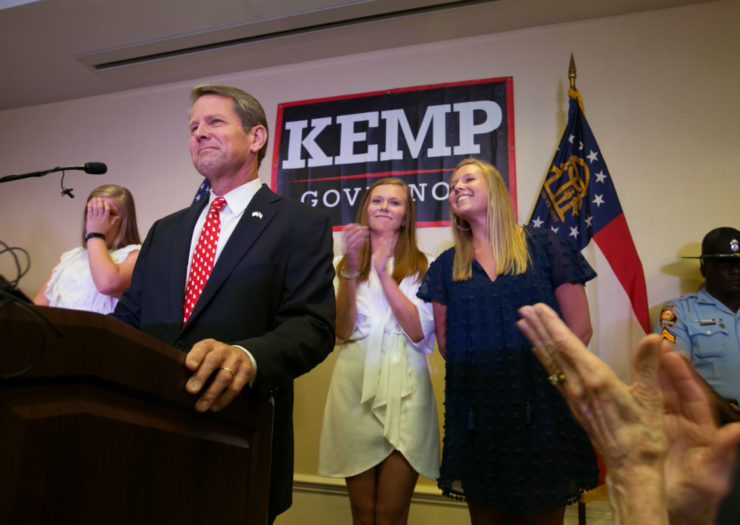 [Photo: Brian Kemp stands at a podium in front of three blonde women and a 
