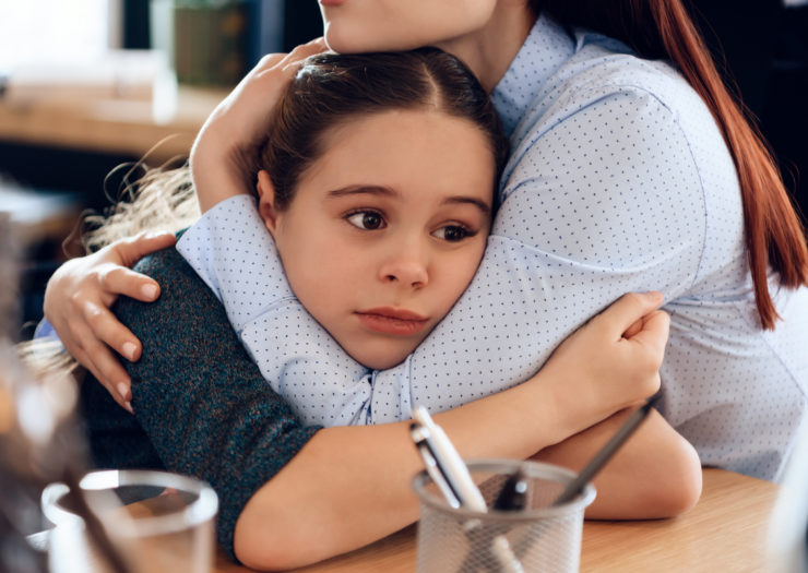 [Photo: Girl with dark hair is hugged by her mother, arms resting on a desk.]