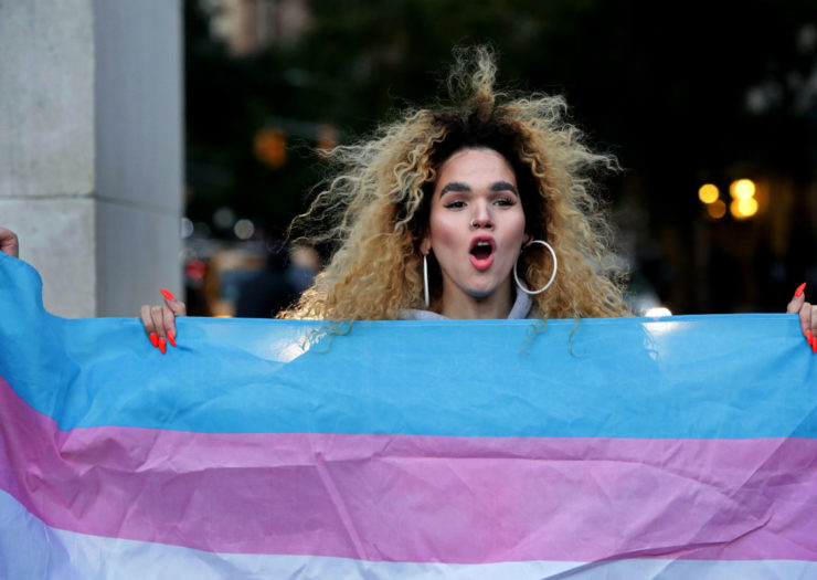[Photo: A trans woman holding up the transgender flag.]