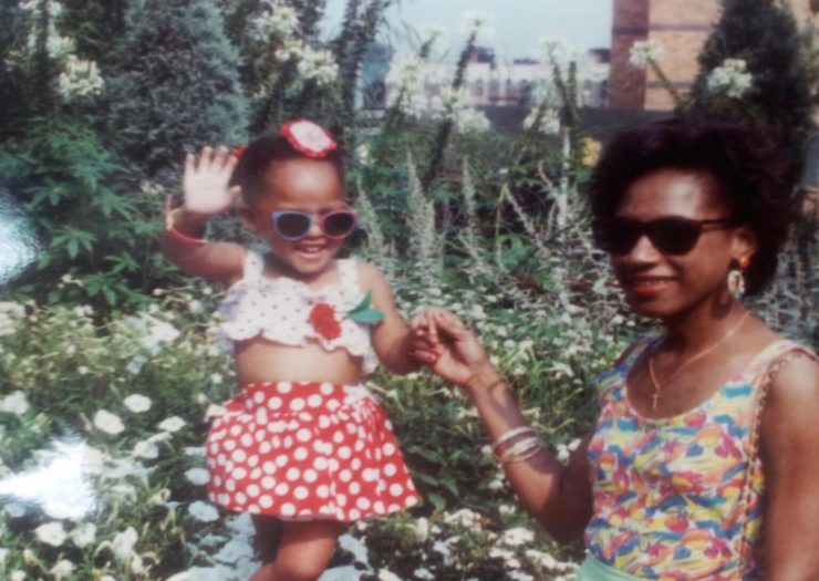 [Tiny Black girl wearing sunglasses and a polka dot dress waves, while a Black woman, also wearing shades, holds her hand and smiles.]