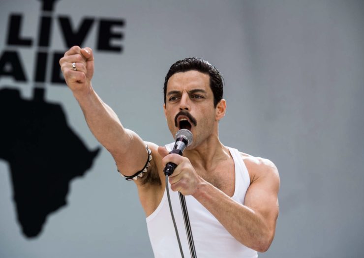 [PHOTO: A dark-haired, hirsute man with a mustache wears a white tank pop and raises a fist while singing or talking into a microphone].