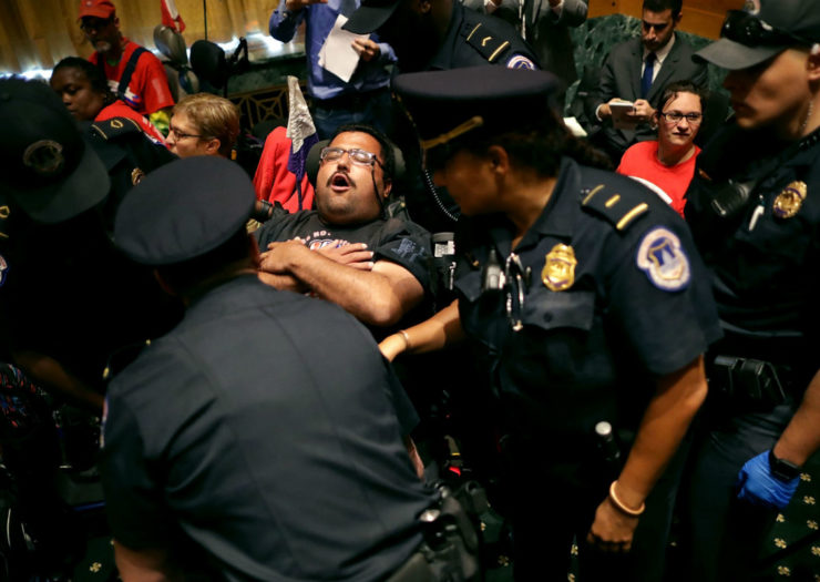 [Photo: A protester in a wheelchair crosses his arms, eyes closed, while Capitol police surround him]