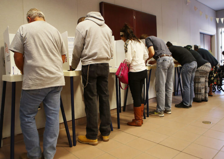 [PHOTO: A line of people facing away from the camera bend over white voting booths as they fill out ballots.]