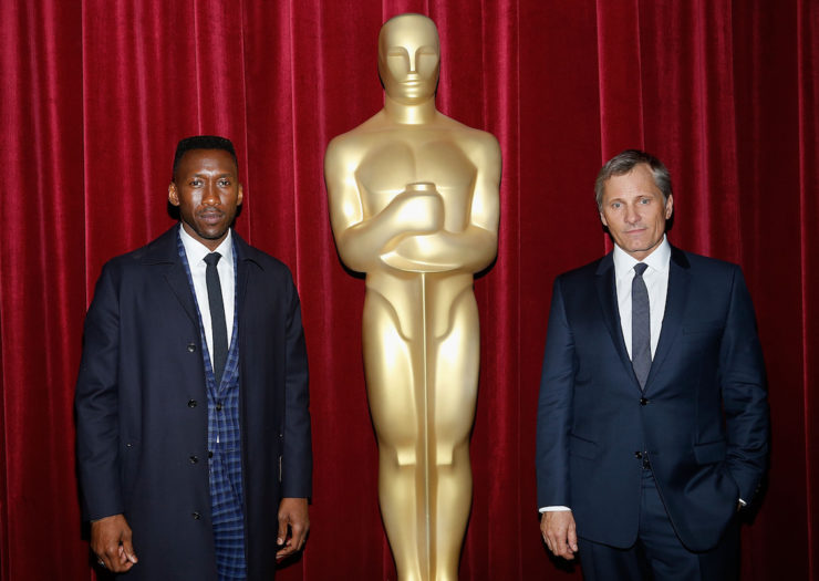 [PHOTO: Black man in a blue plaid suit-overcoat combo and a white man with a touch of gray hair and a much more sober, dark suit flank a giant version of the gold Oscar statue.]