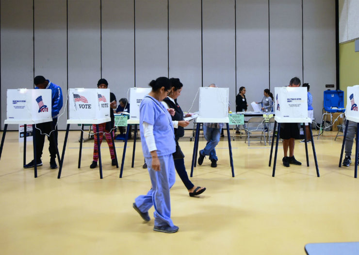[Photo: A woman in scrubs walks past a line of voting booths]
