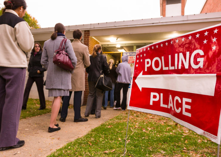 [Photo: People standing in line in front of a polling location.]
