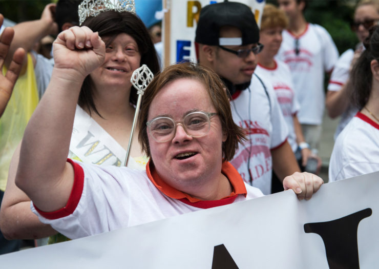 Photo: [A crowd of disabled individuals and allies gather in a rally. At the forefront, a disabled person gestures a power fist as they hold a banner.]