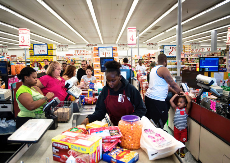 [Photo: Various shoppers wait in line to pay at a grocery store.]
