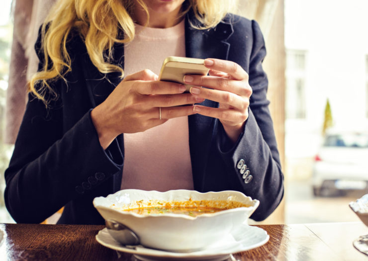 [Photo: Woman holding phone at restaurant table while dining out.]