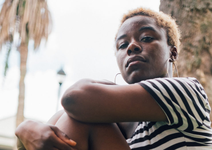 [PHOTO: Serious looking young Black woman with short, blond hair and hoops, sitting outside with palm trees in the background.]