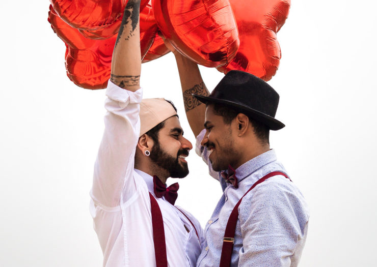 [Photo: Two queer men of color stand face to face with each other as they smile. They're holding red heart balloons as they pose for their engagement photos.]