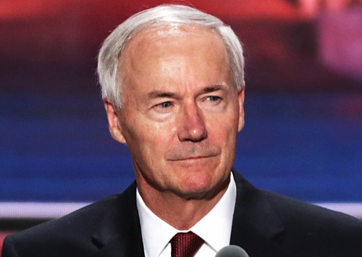 [Photo: Arkansas Governor Asa Hutchinson stands on stage, looking stern, prior to the start of an event.]