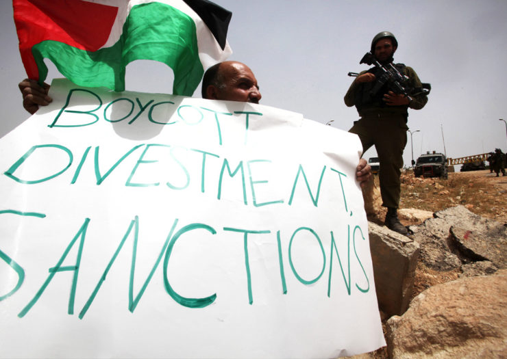 [Photo: A Palestinian holds a placard reading in English ' Boycott divestment, sanctions' as part of a protest.]