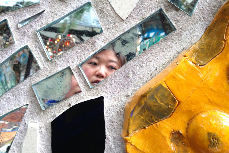 [Photo: An Asian woman takes a photo of herself reflected on the shards of glass that are part of an art composition.}