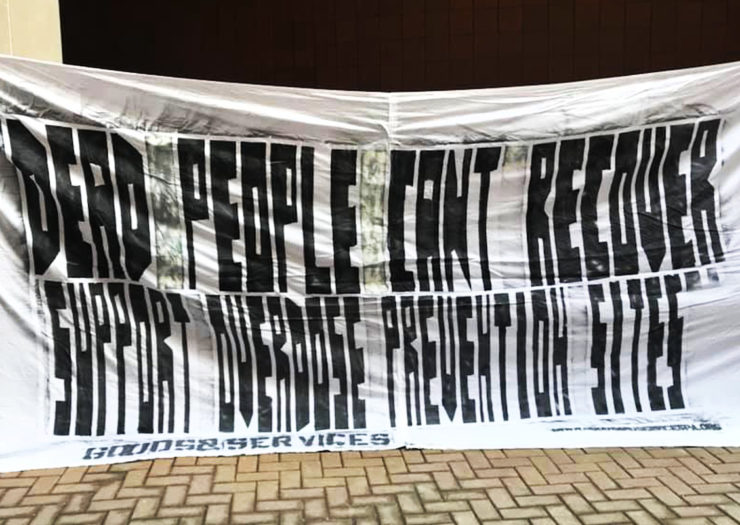[Photo: A white, cloth banner with black text is held up. On it is a message that reads 