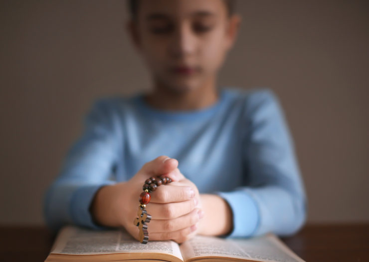 [Photo: Young child praying over a Bible as they hold a rosary.]