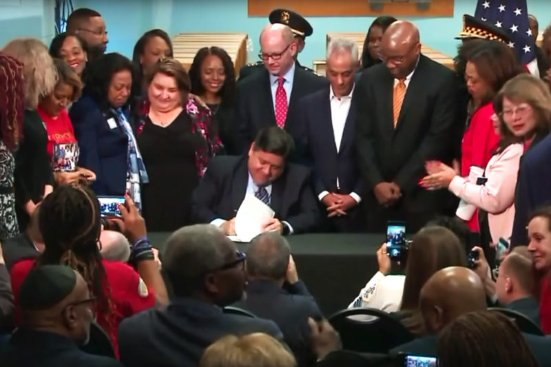 [Photo: Illinois Governor J.B. Pritzker signs legislation in front of the media.]