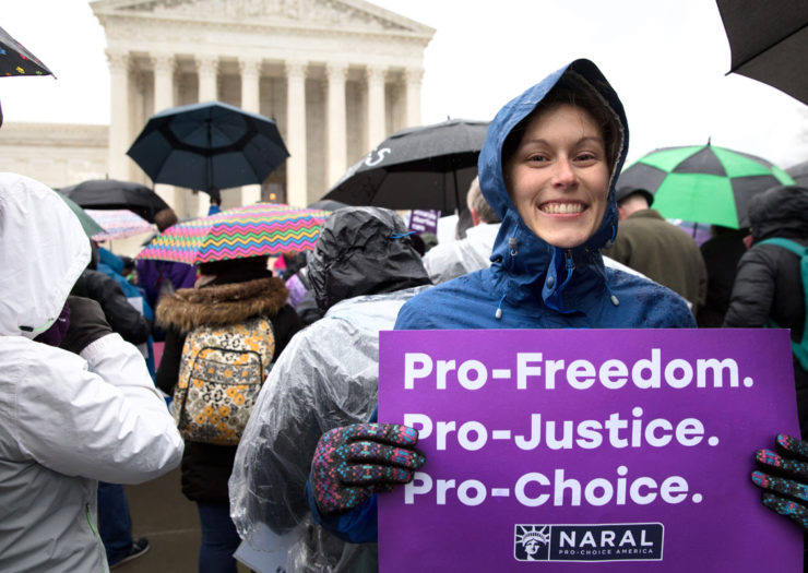[Photo: Young, white woman wears a poncho as she stands in front of the Supreme Court with various others under the rain. She holds a purple sign saying 