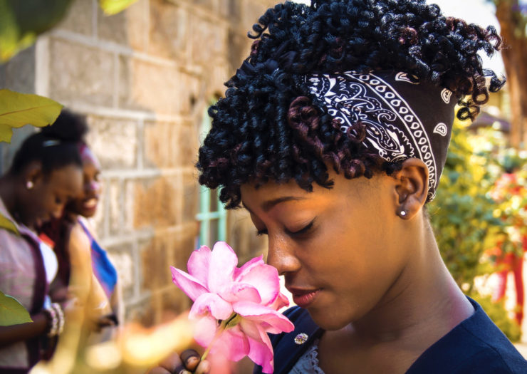 [Photo: A young, black girl smells a flower. Her friends talk amongst themselves in the background.]