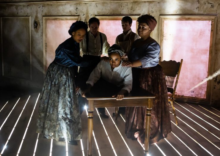 [Photo: A group of actors, among them three Black women, perform a scene from the play Behind the Sheet.]