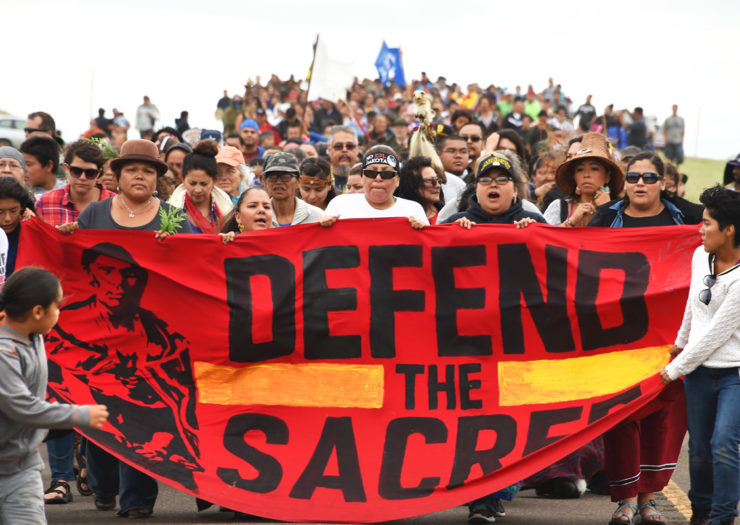 [Photo: Native folks hold a large red banner that reads 