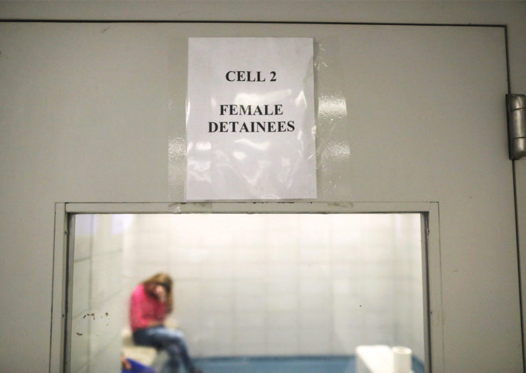 [Photo: A migrant woman is detained in a holding cell. She looks down as she holds her head in her hands.]