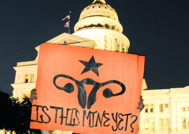[Photo: A person holding a sign in front of the Texas State Capitol. The sign depicts the female reproductive system with a star above it and reads 