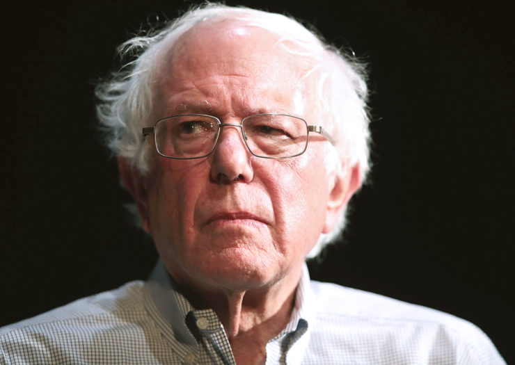 [Photo: Democratic presidential candidate Senator Bernie Sanders looks on during a campaign rally.]